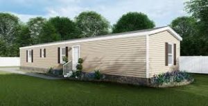 Beige mobile home with white trim and steps, brown shutters, surrounded by grass, small plants, and trees, with a white fence in the background.