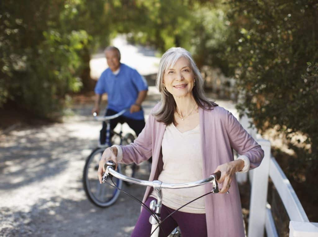 An older woman in a light purple cardigan stands with her bicycle, smiling, at Glenwood Manufactured Home Community, while an older man rides a bike in the background on a sunny path.