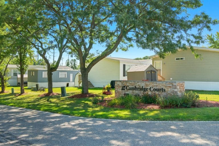 A stone sign reading “Cambridge Courts” stands in front of light-colored manufactured homes, surrounded by trees and well-kept landscaping.