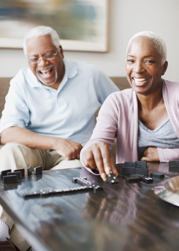 Two older adults smile as they play dominoes together at a table in the well-lit living room of Glenwood Manufactured Home Community.