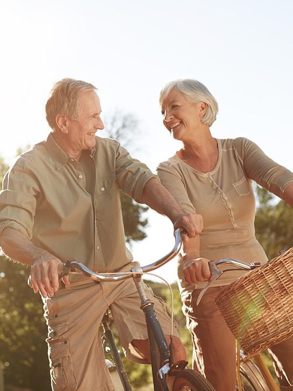 Two older adults ride bicycles outdoors, smiling at each other on a sunny day. A wicker basket is attached to one of the bikes.