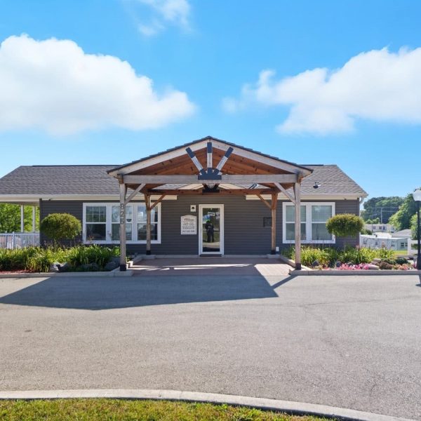 Single-story gray building with a wooden entrance canopy, set within Glenwood Manufactured Home Community, surrounded by landscaping and a parking lot, photographed on a clear sunny day.