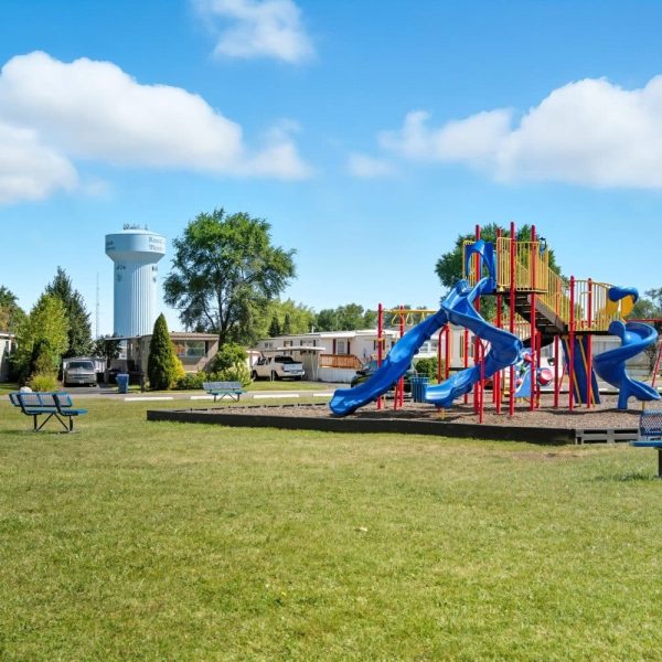 A playground with blue slides and climbing structures sits on a grassy field in Glenwood Manufactured Home Community, with benches nearby and a water tower and houses in the background under a partly cloudy sky.